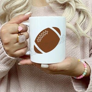 Pode incluir: Caneca de caf&eacute; de cer&acirc;mica branca com um gr&aacute;fico de futebol americano marrom. A caneca est&aacute; sendo segurada por uma pessoa que est&aacute; vestindo um su&eacute;ter rosa.