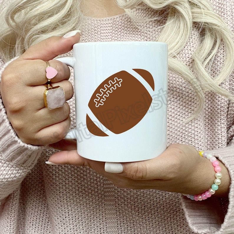 Pode incluir: Caneca de caf&eacute; de cer&acirc;mica branca com um gr&aacute;fico de futebol americano marrom. A caneca est&aacute; sendo segurada por uma pessoa que est&aacute; vestindo um su&eacute;ter rosa.