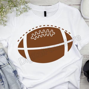 Pode incluir: Camiseta branca com um gr&aacute;fico de futebol americano marrom. A bola de futebol tem costuras brancas e uma linha tracejada ao redor da borda.