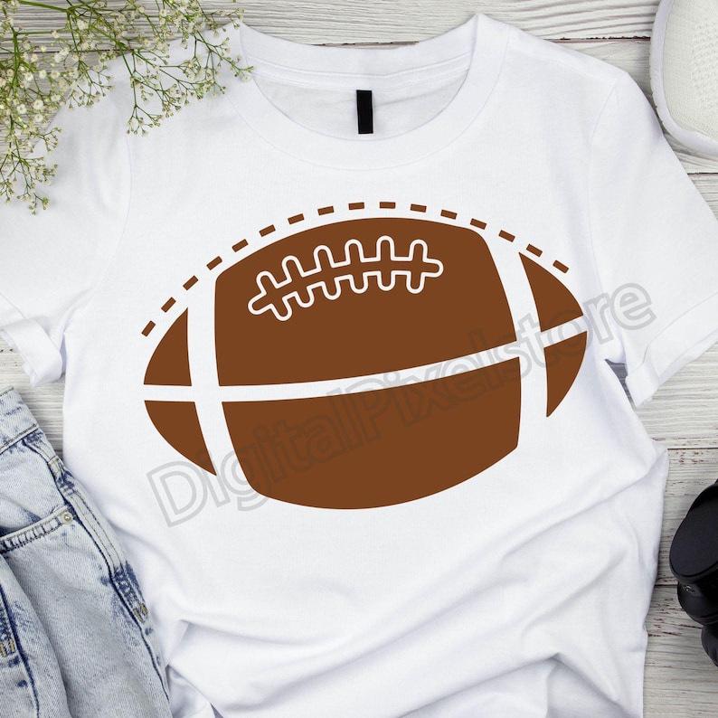 Pode incluir: Camiseta branca com um gr&aacute;fico de futebol americano marrom. A bola de futebol tem costuras brancas e uma linha tracejada ao redor da borda.
