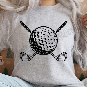 May include: A gray sweatshirt featuring a black graphic of a golf ball with a dimpled surface, crossed by two golf clubs. The design is centered on the shirt, creating a sporty and casual look.