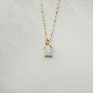 May include: A delicate gold chain necklace with a small oval opal pendant. The opal is white with iridescent flashes of color.