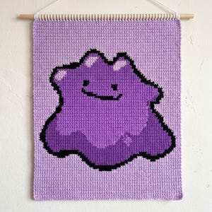 May include: A handmade wall hanging featuring a purple Ditto character from the Pokémon series. The image is a close-up of the crocheted design, with a light purple background and a wooden dowel for hanging.
