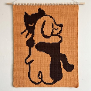 May include: A rectangular crochet wall hanging featuring a dark brown cat and a light tan dog hugging each other. The design is set against a solid mustard yellow background. The piece is suspended from a light wooden dowel with a white string.