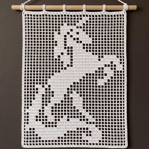 Könnte beinhalten: Ein weißer Wandbehang aus Häkelarbeit mit einem gitterartigen Muster, das die Silhouette eines Einhorns bildet. Das Textilstück ist an einem hellen Holzstab aufgehängt, an dem oben in der Mitte eine weiße Kordel an einem kleinen Haken befestigt ist.