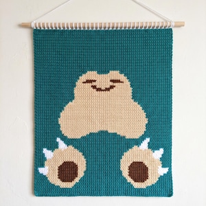Op de afbeelding: Een handgemaakte wandhanger in teal, met een lachend, beige karakter met bruine accenten. De voeten van het personage zijn ook beige met bruine en witte details. De hanger is gemonteerd op een houten deuvel met een wit koord.