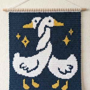 May include: A handmade crochet wall hanging featuring two white geese with yellow beaks and feet against a navy blue background. The design includes yellow star accents and is suspended from a wooden dowel.