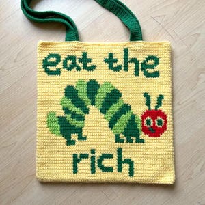 Puede incluir: Bolso tote de crochet amarillo con asas verdes. El bolso presenta un diseño de oruga verde y verde oscuro y las palabras "eat the rich" en letras verdes. La oruga tiene una cabeza roja con una cara sonriente.