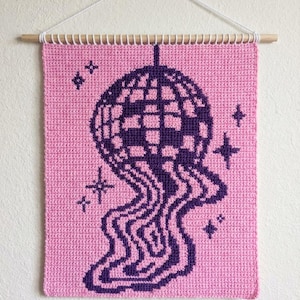 Puede incluir: Colgante de pared rosa y morado con un diseño de bola de discoteca. La bola de discoteca morada tiene un efecto de fusión. El fondo es rosa, con detalles de estrellas moradas. El colgante de pared está hecho de material tejido y se cuelga con una barra de madera, unos 30 cm de ancho.