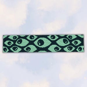 Könnte beinhalten: Ein rechteckiges Strickstirnband mit einem sich wiederholenden Muster aus stilisierten Augen. Das Design besteht aus mintgrünen, mandelförmigen Augen mit dunklen Pupillen auf einem einfarbigen schwarzen Hintergrund, was ein grafisches Motiv ergibt.