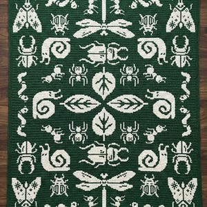 Op de afbeelding: Een vierkant, donkergroen gehaakt kleed met een herhalend patroon van witte insecten, waaronder kevers, libellen en spinnen. Het ontwerp bevat ook bladeren en slakken, wat een op de natuur geïnspireerd thema creëert.