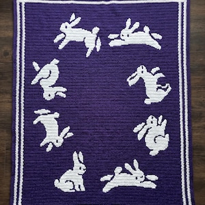 Può includere: Una coperta all'uncinetto fatta a mano di colore viola scuro, con disegni di conigli bianchi. La coperta ha un bordo bianco con un motivo a doppia linea. I conigli sono in varie pose, aggiungendo un tocco giocoso al design.