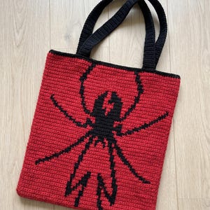 Könnte beinhalten: Eine rechteckige Häkeltasche mit einem leuchtend roten Hintergrund und einer markanten, schwarzen, spinnenartigen Grafik in der Mitte. Die Tasche verfügt über zwei schwarze Häkelgriffe, die an der Oberkante des roten Körpers befestigt sind.