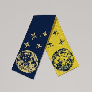 May include: A knitted scarf with a celestial design. One side is navy blue with yellow stars and a moon, the other side is yellow with navy blue stars and a moon. The scarf is approximately 152 cm long.