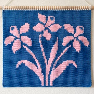 Pode incluir: Um painel de parede de crochê azul com um design floral rosa. As flores e caules são rosa sobre o fundo azul. Pendurado em uma vara de madeira com um cordão branco. Aproximadamente 30 cm quadrados.