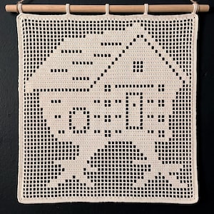 May include: A cream-coloured square crochet wall hanging featuring a filet crochet pattern of a house with a chimney and a circular sun or moon shape below. The textile is suspended from a light-coloured wooden dowel with a thin brown twine hanger.