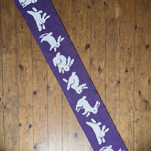May include: A long, rectangular purple scarf featuring a repeating pattern of white crocheted rabbits. The scarf is constructed with a dense, ribbed knit texture, and the white rabbit silhouettes are evenly spaced along the entire length of the fabric.