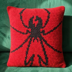 May include: A square crochet throw pillow featuring a vibrant red background with a prominent black spider graphic centered on the front. The textured yarn construction creates a distinct pattern, showcasing the arachnid design clearly.