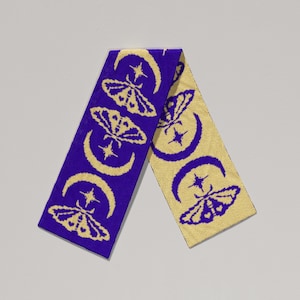May include: A knitted scarf featuring a repeating pattern of moths, crescent moons, and stars. The scarf has a purple background with a gold design. The scarf is made of a soft, knitted material.