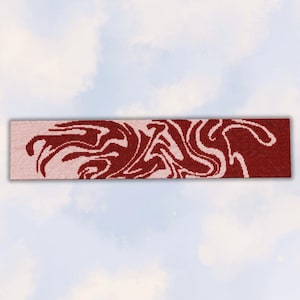 Puede incluir: Una bufanda rectangular de punto con un patrón jaspeado de dos tonos en rojo intenso y rosa claro. El diseño consiste en formas orgánicas fluidas y arremolinadas que abarcan toda la longitud de la tela, creando un gráfico abstracto continuo.