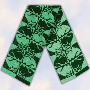 May include: A knitted scarf featuring a repeating grid pattern of pixelated frog faces. The design alternates between dark green and light mint green squares, with each square containing a stylized, simple graphic of a frog with closed eyes.