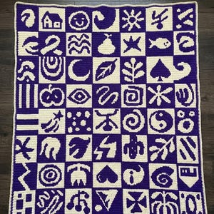 Può includere: Una coperta all'uncinetto fatta a mano con un motivo a griglia di quadrati viola e crema. Ogni quadrato mostra un simbolo unico, tra cui una casa, un pesce e un cuore. La coperta è un oggetto decorativo e accogliente.