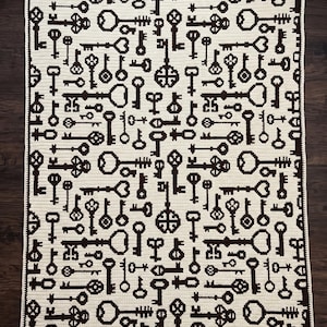 May include: A rectangular crochet blanket featuring a repeating pattern of dark brown keys on a cream-colored background. The design displays various key shapes and sizes oriented in different directions, finished with a dark brown border.