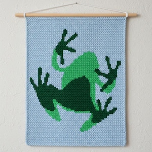 May include: A light blue wall hanging with a green frog design. The frog is depicted in two shades of green, with a detailed, pixelated appearance. The wall hanging is mounted on a wooden dowel with a white cord for hanging.