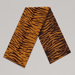May include: A rectangular scarf with a tiger stripe pattern. The scarf features a warm orange background with dark brown stripes. The scarf is made of a soft material and is folded in half.