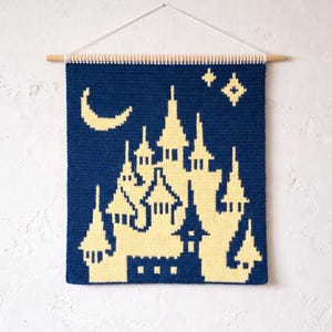 May include: A navy blue wall hanging featuring a yellow castle design, a crescent moon, and stars. The castle has multiple towers and windows. The wall hanging is suspended from a wooden dowel with a white string.