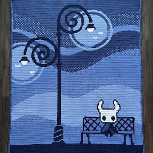 May include: A rectangular crochet blanket featuring a blue-toned scene with a tall dark blue lamppost and two glowing circular lights. A white character with a horned head sits on a dark blue bench in the foreground against a blue background.