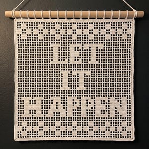 May include: A cream-colored crochet wall hanging featuring a grid pattern with the text LET IT HAPPEN integrated into the design. The textile is suspended from a light-colored wooden dowel with a white string attached for hanging purposes.