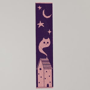 May include: A purple and pink knitted scarf featuring a whimsical design. The design includes a house with smoke, a cat, stars, and a crescent moon. The scarf is rectangular and has a pink border.