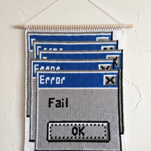 Op de afbeelding: Gehaakte wandhanger met een retro computerfoutmelding ontwerp. Het ontwerp gebruikt blauwe en grijze kleuren met de woorden "Error" en "Fail". Het is gemonteerd op een houten stok.