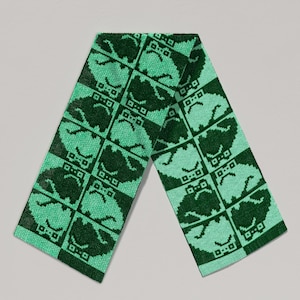 May include: A green knitted scarf with a repeating face pattern. The scarf is rectangular and folded, showcasing the design. The face design is in a pixelated style, adding a unique touch to the accessory. The scarf is a winter accessory.