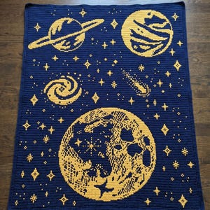 May include: A rectangular crochet blanket featuring a dark blue background with yellow celestial motifs. The design includes a large moon, a planet with rings, a second planet, a spiral galaxy, a comet, and various scattered four-pointed stars.