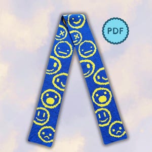 Könnte beinhalten: Ein langer, blauer Strickschal mit einem sich wiederholenden Muster aus gelben, verpixelten Smileys mit verschiedenen Gesichtsausdrücken. Rechts neben dem Schal befindet sich ein hellblaues, rundes Abzeichen mit gewelltem Rand und dem Text PDF.