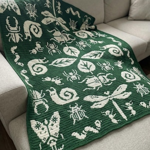 May include: A dark green crochet blanket featuring a repeating cream-colored pattern of various insects, including dragonflies, beetles, snails, and caterpillars, interspersed with leaf motifs across the entire rectangular textile surface.