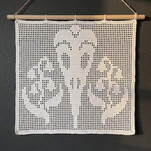May include: A white crochet wall hanging featuring a central bird skull motif flanked by two leafy plant designs. The square textile is suspended from a light-colored wooden dowel with a thin jute twine cord attached for hanging purposes.