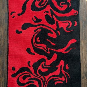 May include: A rectangular, handmade crochet blanket with a bold design. The blanket is split into two sections: a solid red area on the left and a black area on the right. Abstract, swirling shapes in contrasting colours create a dynamic visual effect.
