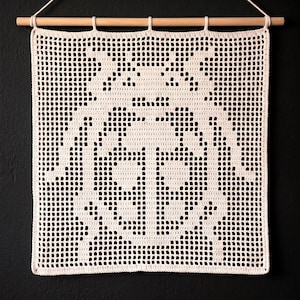 May include: A cream-colored crochet wall hanging featuring a grid-like pattern that forms the silhouette of a beetle. The textile is suspended from a light-colored wooden dowel with a matching string attached for hanging on a wall.