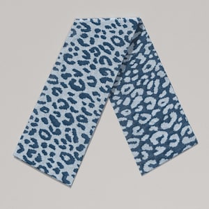 May include: A folded scarf with a blue leopard print pattern. The scarf is made of a soft material and features a repeating pattern of dark blue spots on a light blue background. The scarf is folded in half.