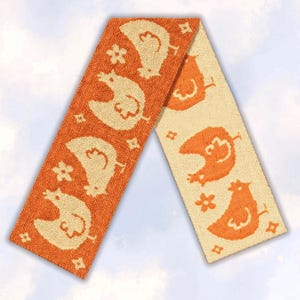 May include: A rectangular woven scarf featuring a repeating pattern of chickens and small four-pointed stars. The scarf is divided into two sections: one with an orange background and cream-coloured motifs, and the other with a cream background and orange motifs.