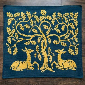 May include: A teal crocheted blanket with a yellow oak tree and two deer beneath. The square blanket has a detailed, textured design. The deer are in a resting position. The blanket is approximately 100 cm x 100 cm.