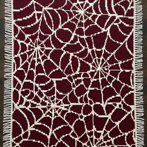 May include: A rectangular crochet blanket featuring a deep burgundy background with an intricate, repeating off-white spider web pattern. The edges of the blanket are finished with a simple border and short, off-white fringe along both sides.