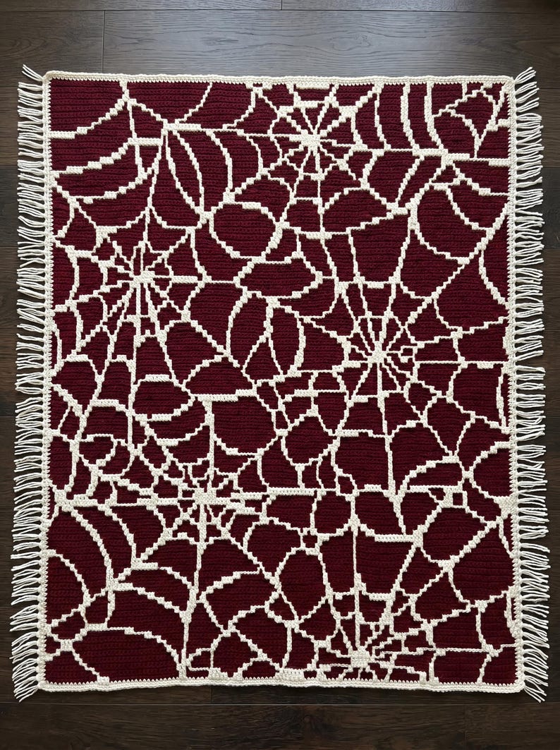 May include: A rectangular crochet blanket featuring a deep burgundy background with an intricate, repeating off-white spider web pattern. The edges of the blanket are finished with a simple border and short, off-white fringe along both sides.