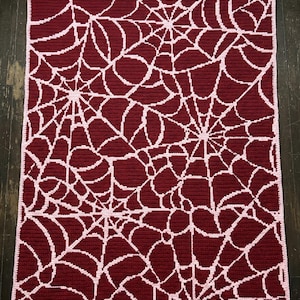 Puede incluir: Una manta rectangular de ganchillo con un patrón repetitivo de telarañas en hilo rosa claro sobre un fondo rojo burdeos intenso. La manta tiene un borde sólido de color rosa claro que enmarca el intrincado diseño en toda la superficie.