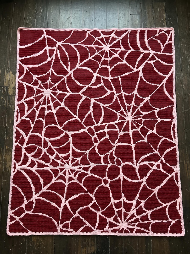 May include: A rectangular crochet blanket featuring a repeating spider web pattern in light pink yarn against a deep burgundy red background. The blanket has a solid light pink border that frames the intricate web design across the entire surface.