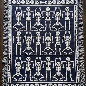 May include: A rectangular navy blue crochet blanket featuring a repeating white pattern of dancing skeletons. The border is decorated with a row of skulls and bones, and the left and right edges are finished with white and navy blue fringe.