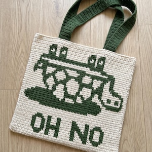 May include: A cream-coloured crochet tote bag featuring a dark green pixelated graphic of a creature emerging from a pool of liquid, with the text "OH NO" printed in dark green block letters below the design and two dark green crochet handles.